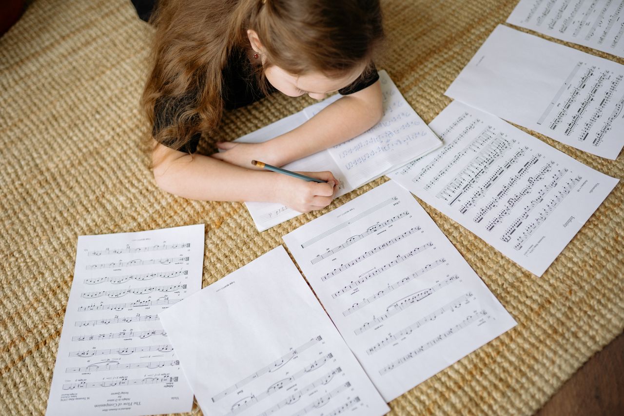 gallery-6 A young girl lying on a carpet writing music notes surrounded by sheets, enjoying a creative moment at home.